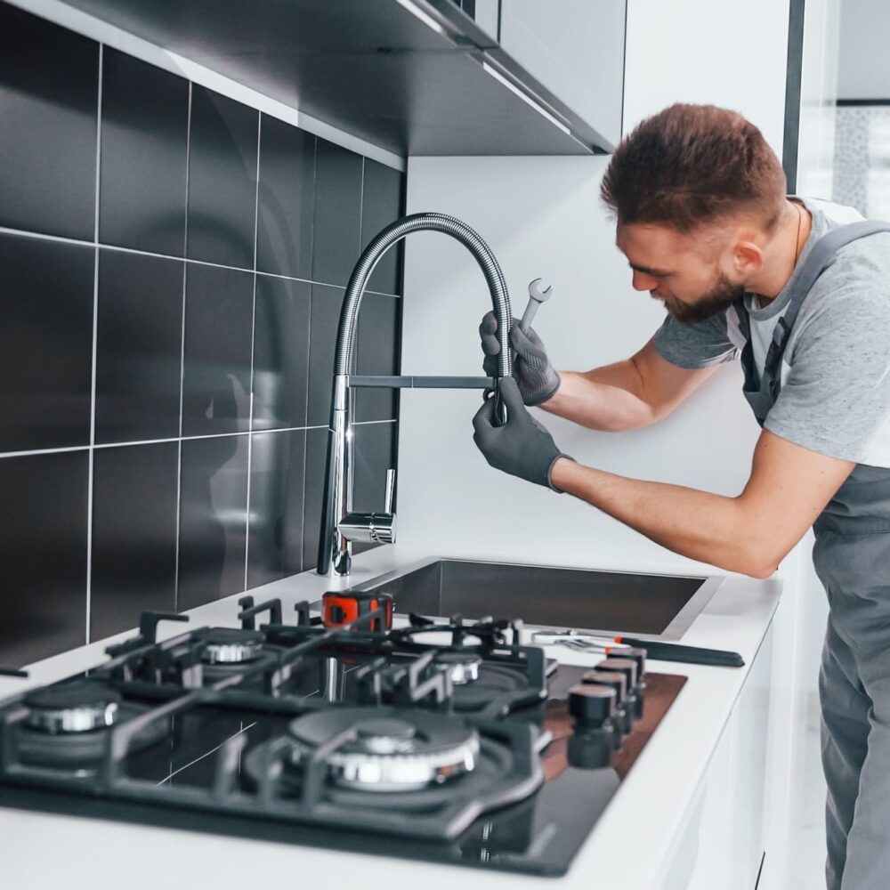 Professional plumber in grey uniform fixing water tap in the kitchen